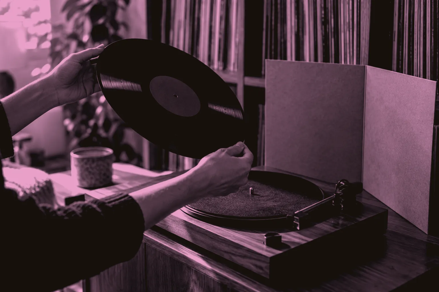 Two hands turn a black LP over above a turntable, beside an open gatefold sleeve and a shelf of upright records.