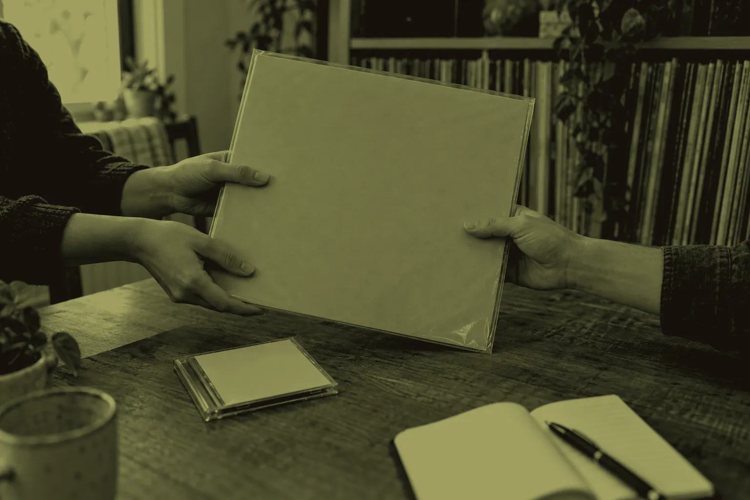 Hands pass a sleeved record across a wooden table beside a CD case and an open notebook.
